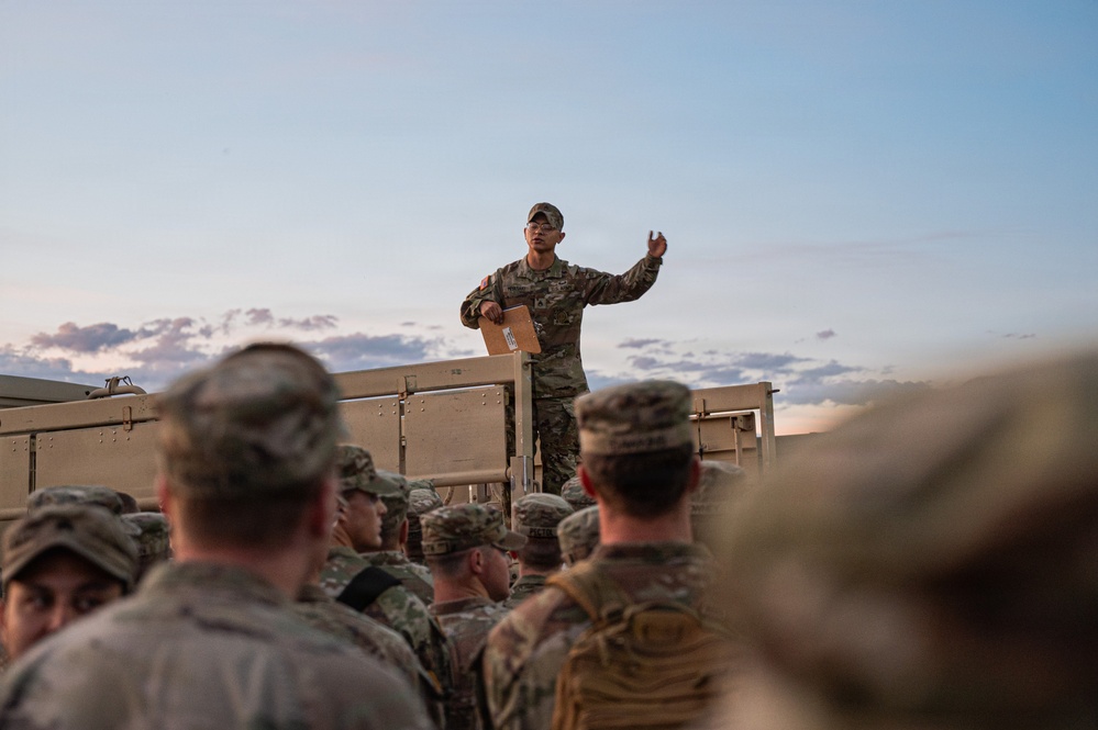 A military service member in US Army OCP (Operational Camouflage Pattern) uniform stands elevated on the bed of a military vehicle, addressing a group of assembled soldiers below. The scene appears to take place at dusk or dawn in an open field or training area. The standing soldier appears to be giving a briefing or issuing instructions while holding what appears to be a document or folder.