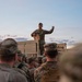 A military service member in US Army OCP (Operational Camouflage Pattern) uniform stands elevated on the bed of a military vehicle, addressing a group of assembled soldiers below. The scene appears to take place at dusk or dawn in an open field or training area. The standing soldier appears to be giving a briefing or issuing instructions while holding what appears to be a document or folder.
