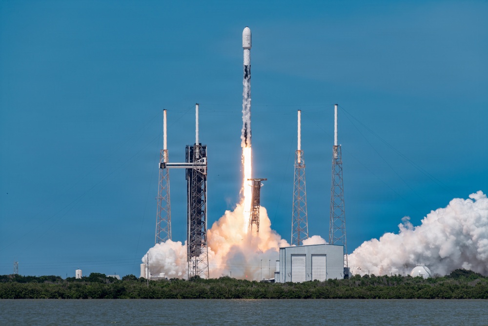 A rocket is captured at the moment of liftoff from a coastal launch pad, with intense engine flames and large billowing clouds of exhaust smoke visible at the base. The rocket, consistent in appearance with a SpaceX Falcon 9 design, is ascending vertically with a white payload fairing at its top. The launch facility features multiple lightning rod towers and support structures surrounding the pad, with a body of water and treeline visible in the foreground and background.