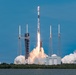 A rocket is captured at the moment of liftoff from a coastal launch pad, with intense engine flames and large billowing clouds of exhaust smoke visible at the base. The rocket, consistent in appearance with a SpaceX Falcon 9 design, is ascending vertically with a white payload fairing at its top. The launch facility features multiple lightning rod towers and support structures surrounding the pad, with a body of water and treeline visible in the foreground and background.