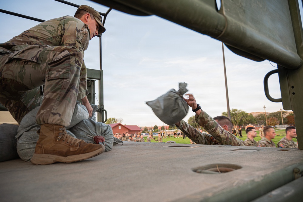 Military personnel in OCP (Operational Camouflage Pattern) uniforms are loading or unloading sandbags from what appears to be the bed of a military vehicle. The photo is taken from a low angle on the vehicle bed, showing one soldier kneeling and handling sandbags while another tosses one. In the background, a large formation of soldiers is visible on a grassy field near red-brick military buildings.