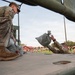 Military personnel in OCP (Operational Camouflage Pattern) uniforms are loading or unloading sandbags from what appears to be the bed of a military vehicle. The photo is taken from a low angle on the vehicle bed, showing one soldier kneeling and handling sandbags while another tosses one. In the background, a large formation of soldiers is visible on a grassy field near red-brick military buildings.
