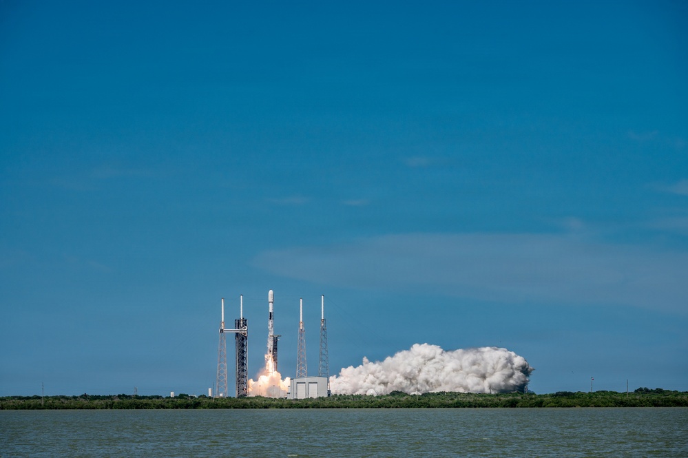 A rocket is captured at the moment of liftoff from a coastal launch pad, with a large plume of white exhaust smoke billowing outward from the base. The launch is photographed from across a body of water, providing a wide, distant perspective of the event. The clear blue sky and calm water create a vivid contrast with the dramatic smoke cloud.