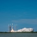 A rocket is captured at the moment of liftoff from a coastal launch pad, with a large plume of white exhaust smoke billowing outward from the base. The launch is photographed from across a body of water, providing a wide, distant perspective of the event. The clear blue sky and calm water create a vivid contrast with the dramatic smoke cloud.