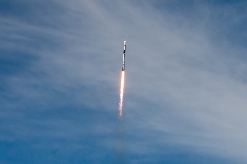 A rocket is captured mid-flight ascending steeply through a partly cloudy blue sky. The rocket is producing a bright orange and white exhaust flame and a visible white contrail trailing below it. The image appears to depict a SpaceX Falcon 9 rocket based on its distinctive white cylindrical body and payload fairing design.