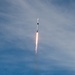 A rocket is captured mid-flight ascending steeply through a partly cloudy blue sky. The rocket is producing a bright orange and white exhaust flame and a visible white contrail trailing below it. The image appears to depict a SpaceX Falcon 9 rocket based on its distinctive white cylindrical body and payload fairing design.
