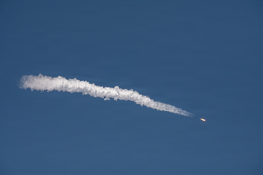 The image shows a rocket or missile in flight against a clear blue sky, leaving a prominent white exhaust/contrail plume behind it. The object is visible on the right side of the frame as a small bright point with an orange-yellow flame, trailing a thick, billowing white smoke trail that extends diagonally across the frame from right to left. The sky is uniformly clear with no clouds, providing strong contrast against the white plume.