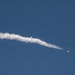 The image shows a rocket or missile in flight against a clear blue sky, leaving a prominent white exhaust/contrail plume behind it. The object is visible on the right side of the frame as a small bright point with an orange-yellow flame, trailing a thick, billowing white smoke trail that extends diagonally across the frame from right to left. The sky is uniformly clear with no clouds, providing strong contrast against the white plume.