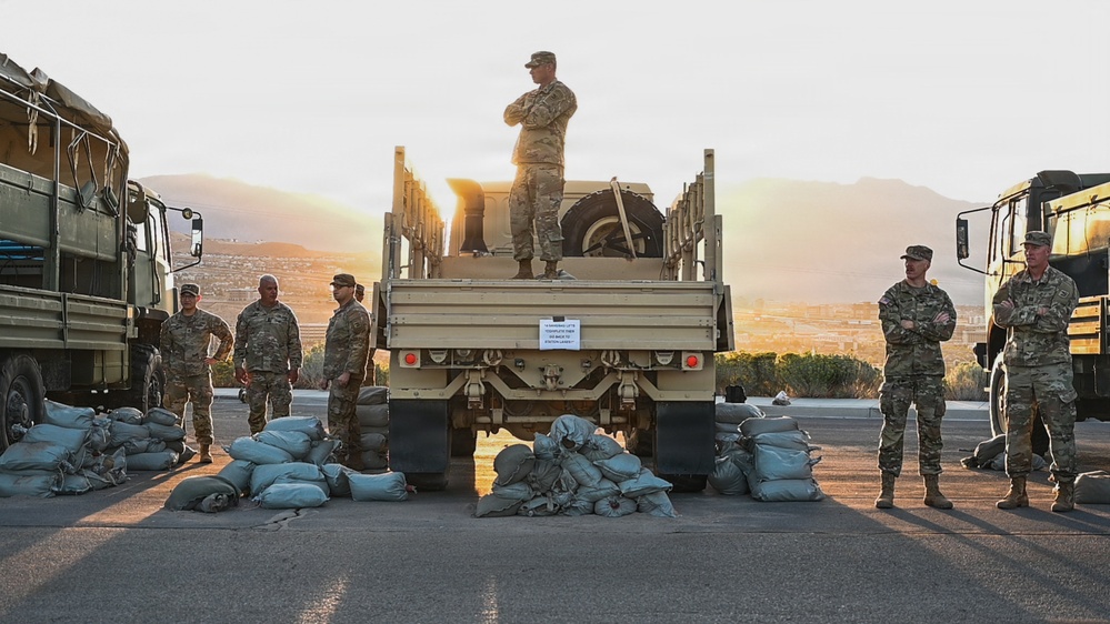 A group of military personnel in camouflage uniforms are assembled around military trucks on a paved surface at dawn or dusk, with mountains and a city visible in the background. Numerous sandbags are arranged on the ground in front of the vehicles. One soldier stands atop a military flatbed truck with arms crossed, appearing to oversee the operation.
