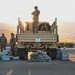 A group of military personnel in camouflage uniforms are assembled around military trucks on a paved surface at dawn or dusk, with mountains and a city visible in the background. Numerous sandbags are arranged on the ground in front of the vehicles. One soldier stands atop a military flatbed truck with arms crossed, appearing to oversee the operation.