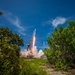 A rocket is captured mid-launch from a ground-level perspective framed by dense green vegetation on both sides. The rocket, consistent in appearance with a SpaceX Falcon 9, is ascending with a bright orange flame and billowing white exhaust plume visible at the base. The image was taken during daytime under a vivid blue sky with scattered cirrus clouds.