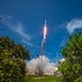 A rocket is captured mid-launch, ascending steeply into a clear blue sky with visible engine flames and exhaust plumes. The photo is taken from a low angle through surrounding vegetation, providing a dramatic ground-level perspective of the launch. The launch infrastructure, including a lightning rod tower and support structures, is visible at the base.