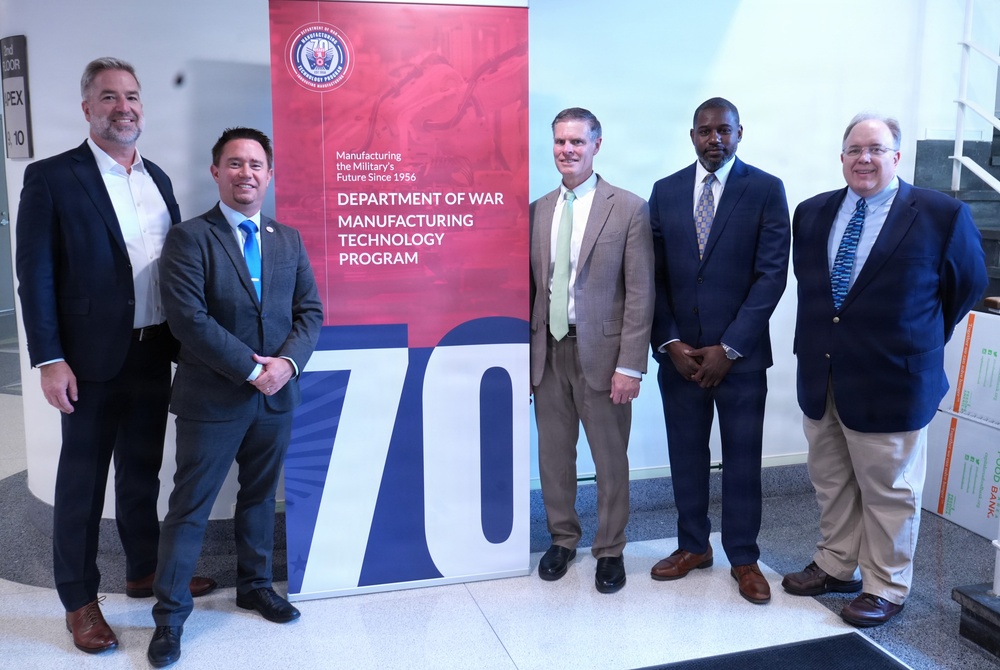 Five men in business attire pose for a group photo in front of a promotional banner for the Department of War Manufacturing Technology Program, celebrating its 70th anniversary since 1956. The setting appears to be an indoor facility or conference space. The occasion appears to be an official or commemorative event related to the program's milestone anniversary.
