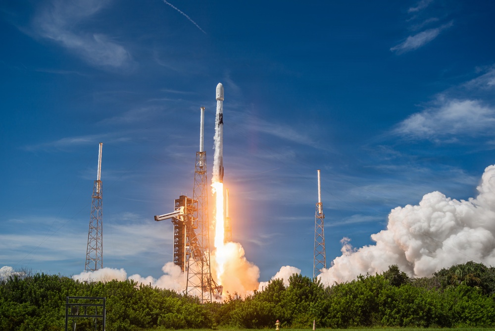 A rocket is captured at the moment of liftoff from a launch pad, surrounded by intense flames, exhaust plumes, and billowing white smoke clouds. The rocket appears to be a SpaceX Falcon 9, identifiable by its white cylindrical body and characteristic fairing shape. The launch is taking place in a coastal, vegetated area under a partly cloudy blue sky.
