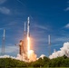 A rocket is captured at the moment of liftoff from a launch pad, surrounded by intense flames, exhaust plumes, and billowing white smoke clouds. The rocket appears to be a SpaceX Falcon 9, identifiable by its white cylindrical body and characteristic fairing shape. The launch is taking place in a coastal, vegetated area under a partly cloudy blue sky.