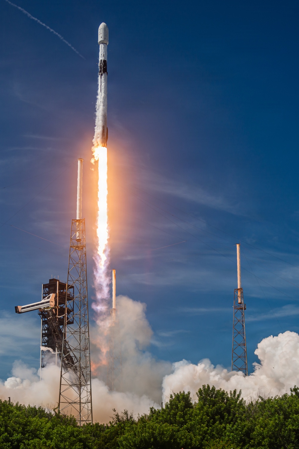 A rocket is captured mid-launch, ascending vertically with a bright plume of fire and billowing white exhaust clouds at its base. The launch infrastructure, including a steel launch tower with lightning rods, is visible in the foreground surrounded by green vegetation. The rocket's white body and payload fairing are clearly visible against a vivid blue sky.