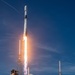 A rocket is captured mid-launch, ascending vertically with a bright plume of fire and billowing white exhaust clouds at its base. The launch infrastructure, including a steel launch tower with lightning rods, is visible in the foreground surrounded by green vegetation. The rocket's white body and payload fairing are clearly visible against a vivid blue sky.
