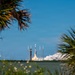A rocket is captured at the moment of liftoff from a coastal launch pad, photographed from across a body of water through framing palm and scrub vegetation. Large plumes of white exhaust smoke billow around the base of the rocket as it begins its ascent. The rocket's slender white body and the surrounding launch infrastructure are clearly visible against a bright blue sky.