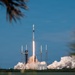 A rocket is captured mid-launch from a coastal launch pad, producing a bright plume of fire and large billowing clouds of exhaust smoke. The rocket, consistent in design with a SpaceX Falcon 9, is ascending vertically against a clear blue sky. The scene is framed by tropical palm fronds in the foreground, suggesting a Florida launch site such as Cape Canaveral.