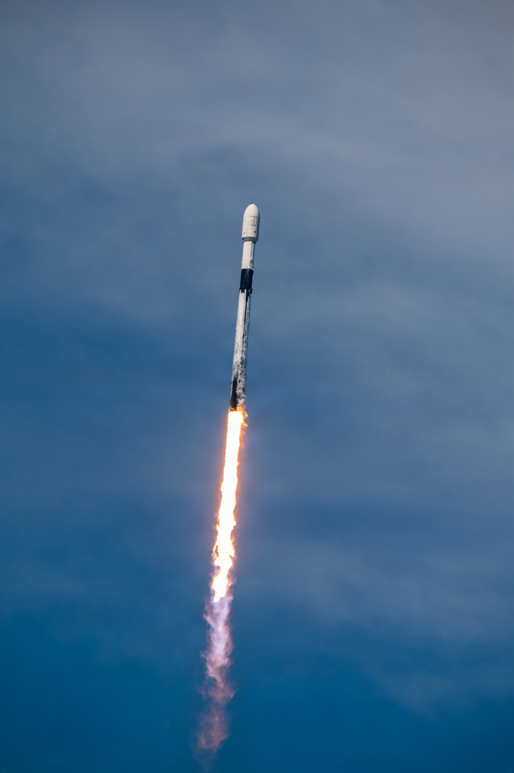 A rocket is captured mid-ascent against a blue, partly cloudy sky. The vehicle displays a white cylindrical body with a dark interstage band, topped by a payload fairing, with a bright orange-white exhaust plume and white smoke trail visible below the engines. The rocket appears to be climbing steeply and has already gained significant altitude.