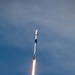 A rocket is captured mid-ascent against a blue, partly cloudy sky. The vehicle displays a white cylindrical body with a dark interstage band, topped by a payload fairing, with a bright orange-white exhaust plume and white smoke trail visible below the engines. The rocket appears to be climbing steeply and has already gained significant altitude.