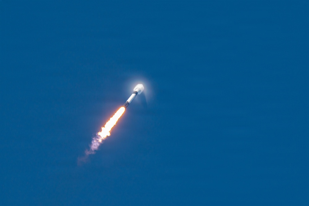 A rocket is captured mid-flight ascending through a clear blue sky at an angle. Bright orange and yellow engine flames are visible at the base, accompanied by a white exhaust plume trailing behind. A condensation or shock wave halo is visible near the nose cone, suggesting the rocket is traveling at high velocity.