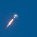 A rocket is captured mid-flight ascending through a clear blue sky at an angle. Bright orange and yellow engine flames are visible at the base, accompanied by a white exhaust plume trailing behind. A condensation or shock wave halo is visible near the nose cone, suggesting the rocket is traveling at high velocity.