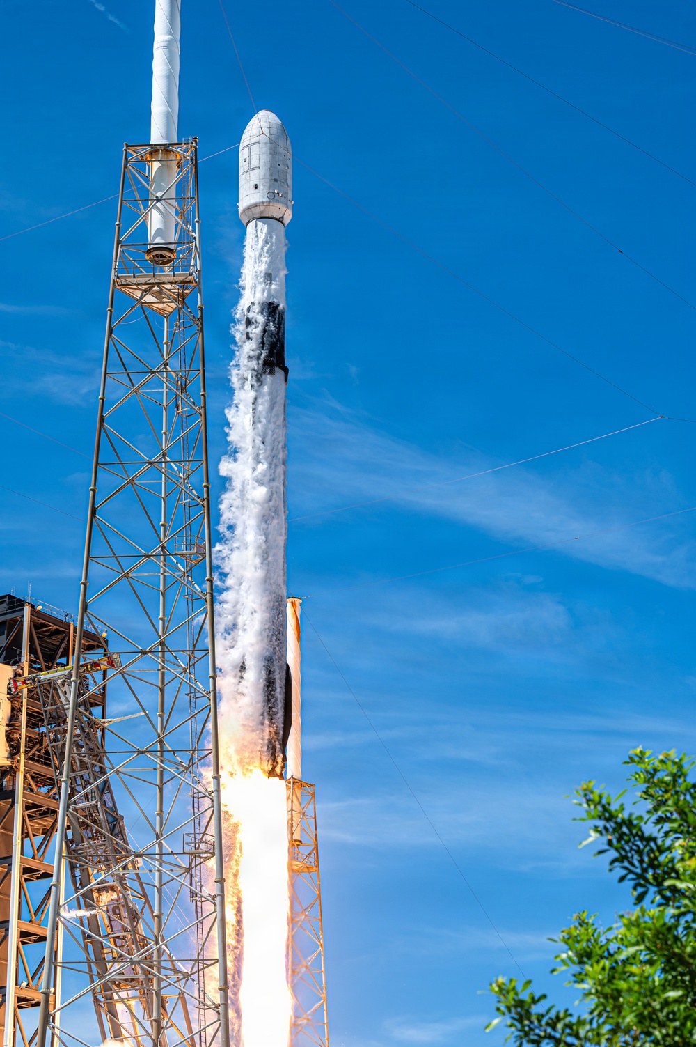 A rocket is captured at the moment of liftoff from a launch pad, with intense flames and white steam/exhaust billowing around the base. The rocket, identifiable by its white cylindrical body and payload fairing at the top, is ascending past the launch support structure under a clear blue sky. The image is taken from a low angle, emphasizing the scale and power of the launch.