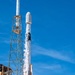 A rocket is captured at the moment of liftoff from a launch pad, with intense flames and white steam/exhaust billowing around the base. The rocket, identifiable by its white cylindrical body and payload fairing at the top, is ascending past the launch support structure under a clear blue sky. The image is taken from a low angle, emphasizing the scale and power of the launch.