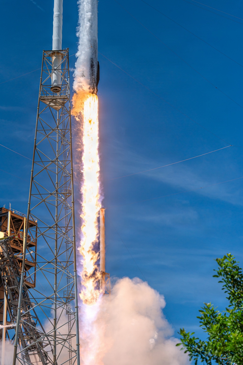 A rocket is captured at the moment of liftoff from a launch pad, with intense engine flames and exhaust plumes visible beneath the vehicle. The rocket is ascending past a tall steel launch tower structure against a clear blue sky. The image captures the early seconds of launch with dramatic fire, smoke, and steam clouds billowing at the base.