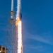 A rocket is captured at the moment of liftoff from a launch pad, with intense engine flames and exhaust plumes visible beneath the vehicle. The rocket is ascending past a tall steel launch tower structure against a clear blue sky. The image captures the early seconds of launch with dramatic fire, smoke, and steam clouds billowing at the base.