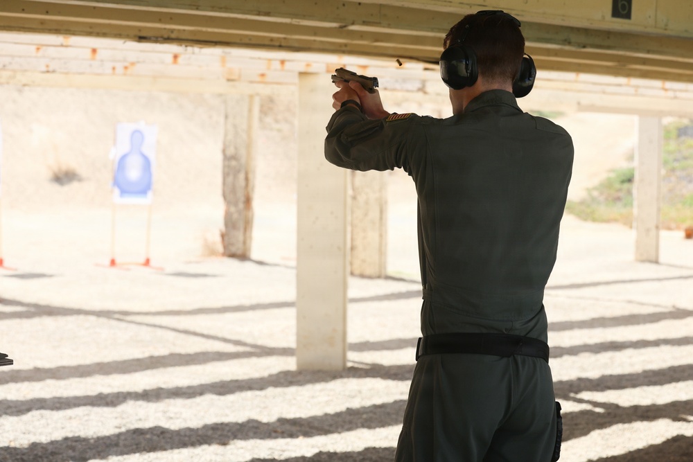 A person is practicing at an outdoor shooting range, aiming a handgun at a blue silhouette target downrange. The shooter is wearing olive/dark green clothing, a black belt, and black ear protection (earmuffs). The range appears to be a covered, open-air facility with concrete support columns and a gravel floor.