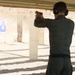 A person is practicing at an outdoor shooting range, aiming a handgun at a blue silhouette target downrange. The shooter is wearing olive/dark green clothing, a black belt, and black ear protection (earmuffs). The range appears to be a covered, open-air facility with concrete support columns and a gravel floor.