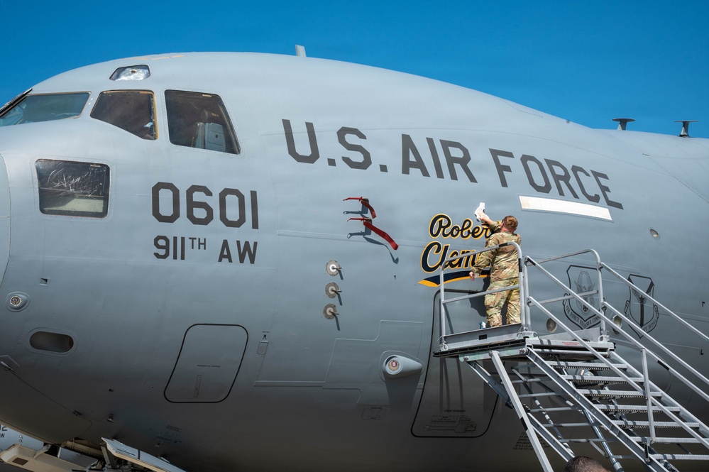 A U.S. Air Force military transport aircraft, tail number 0601 assigned to the 911th Airlift Wing, is being decorated with nose art bearing the name 'Roberto Clemente'. A service member in camouflage uniform is standing on a mobile staircase applying or unveiling the artwork on the fuselage. The image is taken on a clear, sunny day on what appears to be a flight line or tarmac.