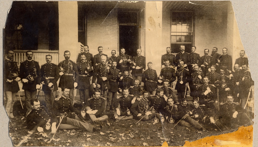 A large group of military officers posed formally in front of a brick building, likely dating from the American Civil War era or late 19th century based on uniform styles. The men are arranged in multiple rows — some standing, some seated, and some reclining on the ground — in a classic formal group portrait composition. The photograph shows significant age-related deterioration including tears, creases, and foxing.
