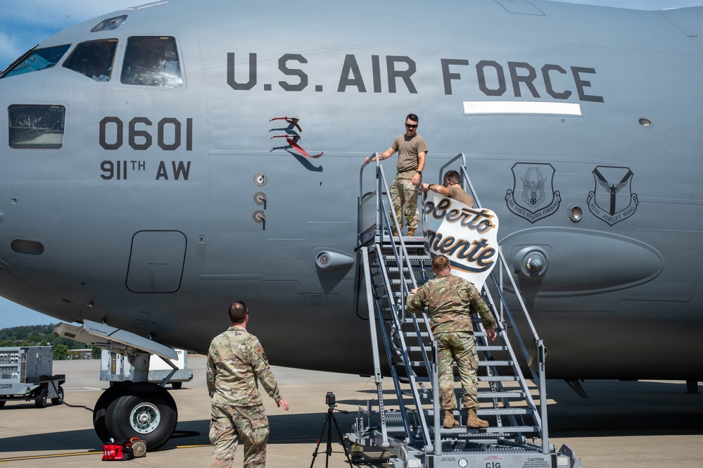U.S. Air Force personnel from the 911th Airlift Wing are working on or near a military transport aircraft (tail number 0601) on a flight line. They appear to be installing or positioning a sign reading 'Roberto Clemente' on the aircraft's boarding stairs. This appears to be a naming ceremony or commemorative event honoring the legendary baseball player Roberto Clemente.