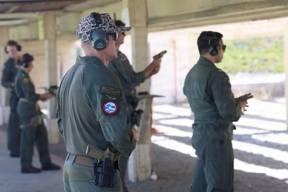 A group of military personnel in olive green flight suits are participating in a firearms qualification or training exercise at an outdoor shooting range. Multiple individuals are visible, some actively aiming pistols downrange while others observe or wait their turn. All participants are wearing hearing protection, consistent with standard range safety protocols.