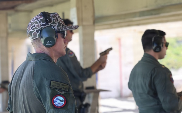 A group of military personnel in olive green flight suits are participating in a firearms qualification or training exercise at an outdoor shooting range. Multiple individuals are visible, some actively aiming pistols downrange while others observe or wait their turn. All participants are wearing hearing protection, consistent with standard range safety protocols.
