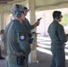 A group of military personnel in olive green flight suits are participating in a firearms qualification or training exercise at an outdoor shooting range. Multiple individuals are visible, some actively aiming pistols downrange while others observe or wait their turn. All participants are wearing hearing protection, consistent with standard range safety protocols.