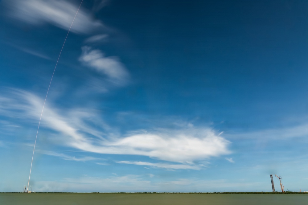 A long-exposure photograph captures a rocket launch trail streaking diagonally across a vivid blue sky, originating from a launch pad visible on the far left horizon. The extended exposure has blurred moving clouds into soft, wispy streaks while preserving the sharp, bright trajectory line of the ascending rocket. Industrial structures, likely a launch tower and crane, are visible on the right side of the horizon.