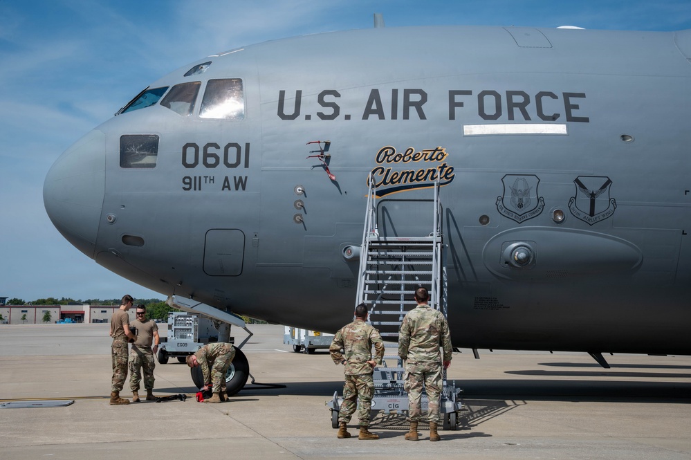A U.S. Air Force aircraft, tail number 0601, assigned to the 911th Airlift Wing, is shown on a flight line with nose art honoring baseball legend Roberto Clemente. Several airmen in camouflage uniforms are gathered around the front of the aircraft, performing ground operations. The image was taken outdoors under partly cloudy skies on what appears to be a military airfield.