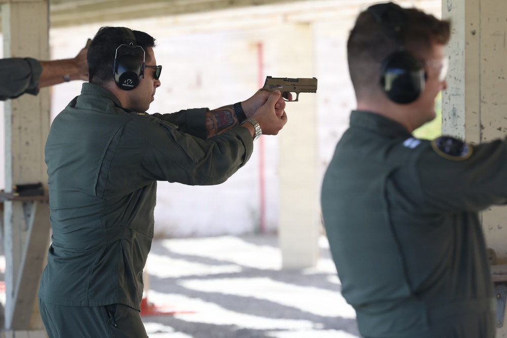 Two individuals wearing olive/sage green flight suits are at an outdoor shooting range. The person in the foreground is actively aiming a tan/FDE-colored semi-automatic pistol in a two-handed grip stance. Both individuals are wearing hearing protection earmuffs and protective eyewear.