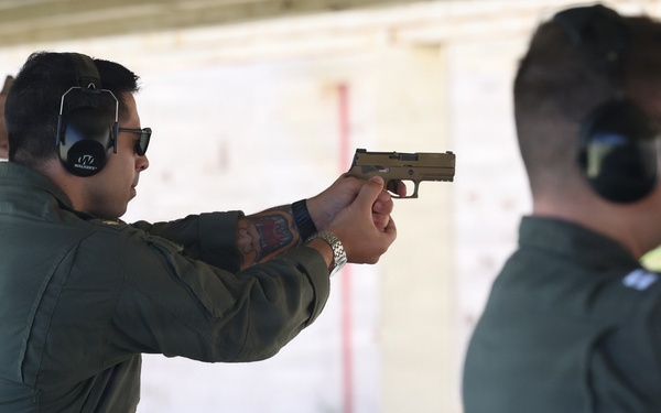 Two individuals wearing olive/sage green flight suits are at an outdoor shooting range. The person in the foreground is actively aiming a tan/FDE-colored semi-automatic pistol in a two-handed grip stance. Both individuals are wearing hearing protection earmuffs and protective eyewear.