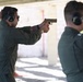 Two individuals wearing olive/sage green flight suits are at an outdoor shooting range. The person in the foreground is actively aiming a tan/FDE-colored semi-automatic pistol in a two-handed grip stance. Both individuals are wearing hearing protection earmuffs and protective eyewear.