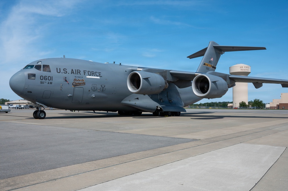 A U.S. Air Force C-17 Globemaster III military transport aircraft, tail number 0601, assigned to the 911th Airlift Wing, is parked on a tarmac at an Air Force Reserve installation. The aircraft bears a special nose art dedication reading 'Roberto Clemente.' A water tower labeled 'Air Force Reserve' is visible in the background.