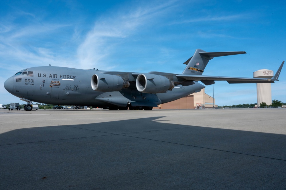 A U.S. Air Force C-17 Globemaster III military transport aircraft is parked on a tarmac at what appears to be an Air Force Reserve base. The aircraft bears the tail number 0601, is assigned to the 911th Airlift Wing, and has a nose art inscription reading 'Roberto Clemente'. A water tower labeled 'Air Force Reserve' is visible in the background.