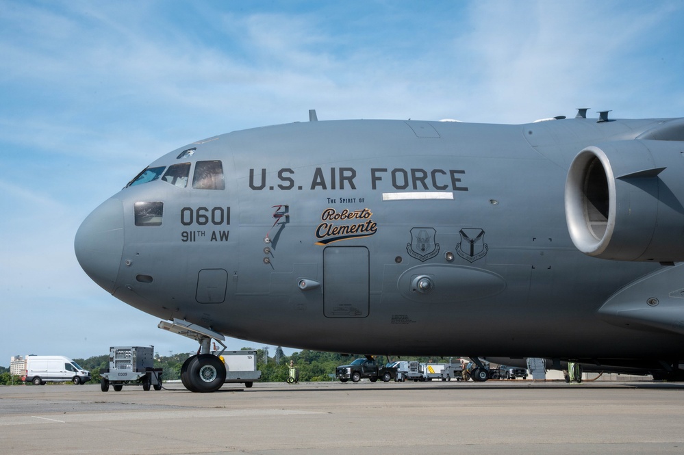 A U.S. Air Force C-17 Globemaster III military transport aircraft is parked on a tarmac. The aircraft bears the nose art designation 'The Spirit of Roberto Clemente' along with tail number 0601 and unit marking 911th AW (Airlift Wing). Ground support equipment and vehicles are visible in the background.