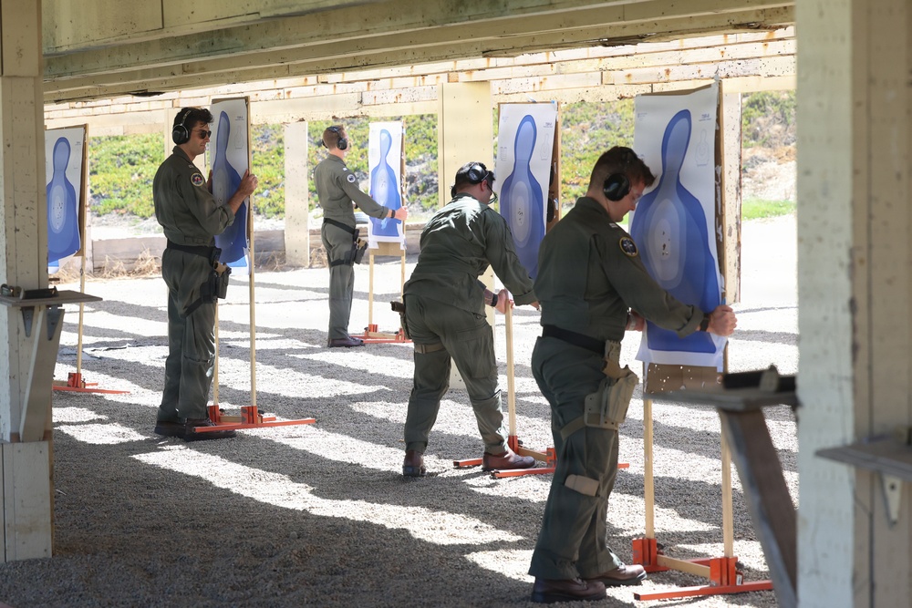Four individuals in matching olive/green military or law enforcement flight suits are setting up or replacing paper shooting targets at an outdoor firing range. They are all wearing ear protection (hearing muffs) and appear to be attaching blue silhouette targets to wooden stands with orange bases. The setting is a covered shooting range with a gravel floor and natural vegetation visible in the background.