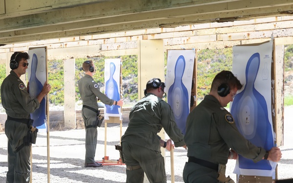 Four individuals in matching olive/green military or law enforcement flight suits are setting up or replacing paper shooting targets at an outdoor firing range. They are all wearing ear protection (hearing muffs) and appear to be attaching blue silhouette targets to wooden stands with orange bases. The setting is a covered shooting range with a gravel floor and natural vegetation visible in the background.