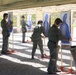 Four individuals in matching olive/green military or law enforcement flight suits are setting up or replacing paper shooting targets at an outdoor firing range. They are all wearing ear protection (hearing muffs) and appear to be attaching blue silhouette targets to wooden stands with orange bases. The setting is a covered shooting range with a gravel floor and natural vegetation visible in the background.